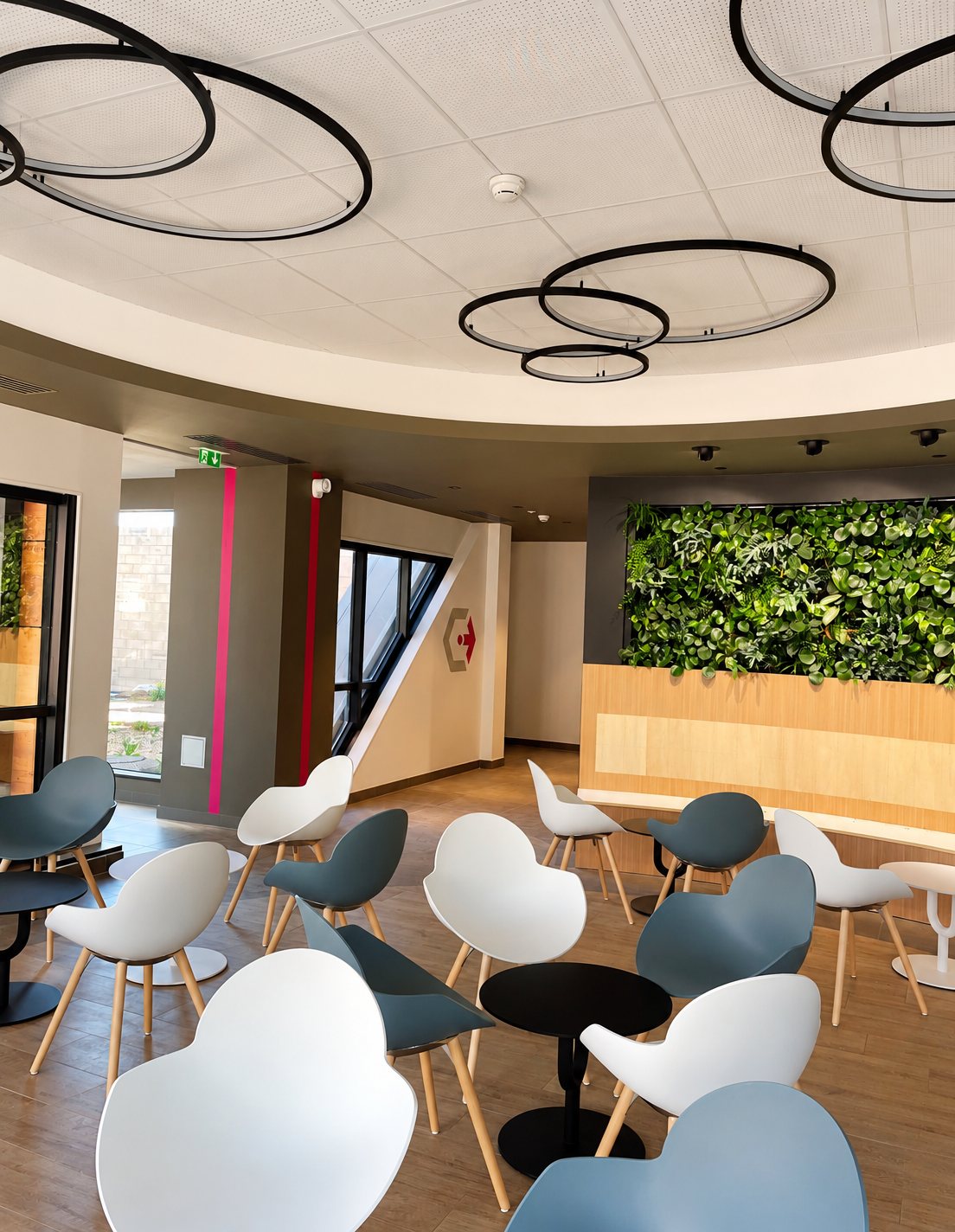 Un espace d'accueil moderne avec des chaises blanches et bleu-gris autour de petites tables rondes, un mur végétal et des suspensions circulaires noires au plafond.
