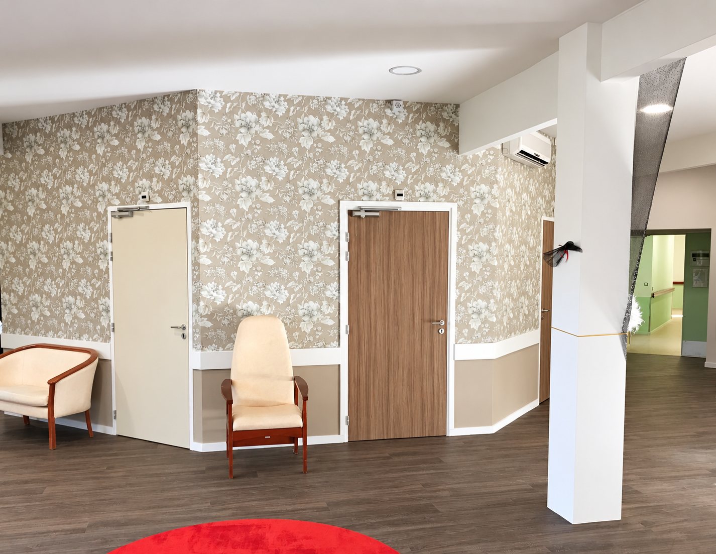 Pièce commune rénovée aux murs tapissés de papier peint à motifs floraux beiges, avec deux portes, deux fauteuils crème, un sol sombre et un tapis rouge au premier plan.