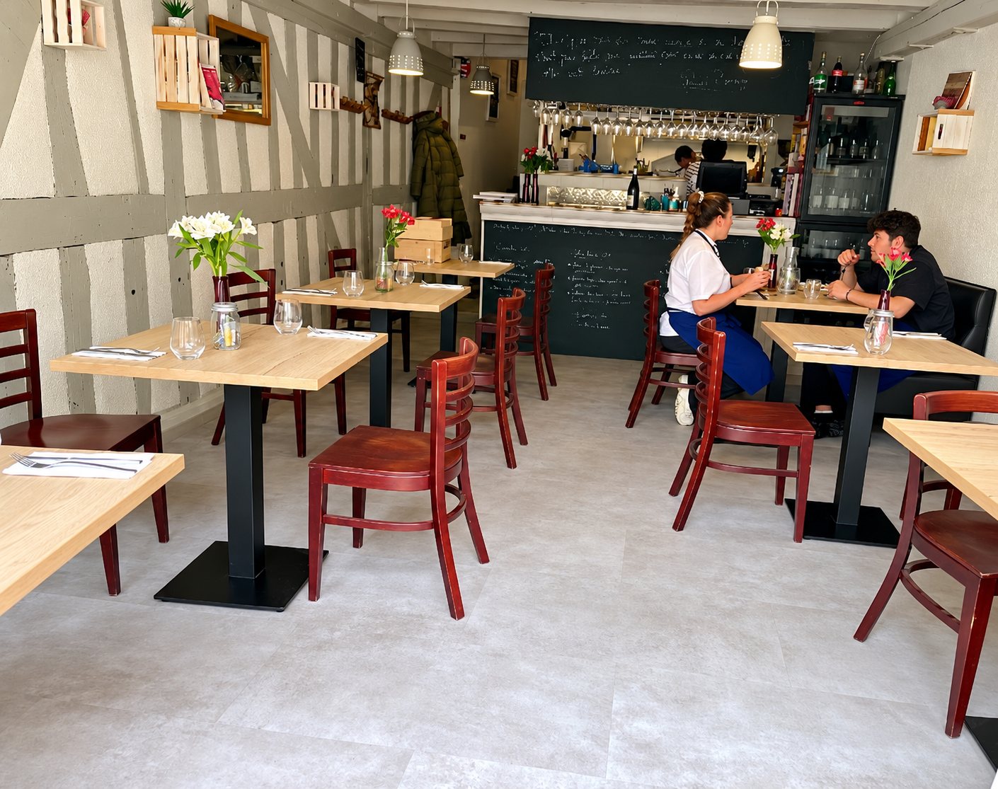 Intérieur d'un petit restaurant aux murs à colombages, avec tables en bois clair, chaises rouges, bouquets de fleurs et deux personnes attablées au fond près du bar.