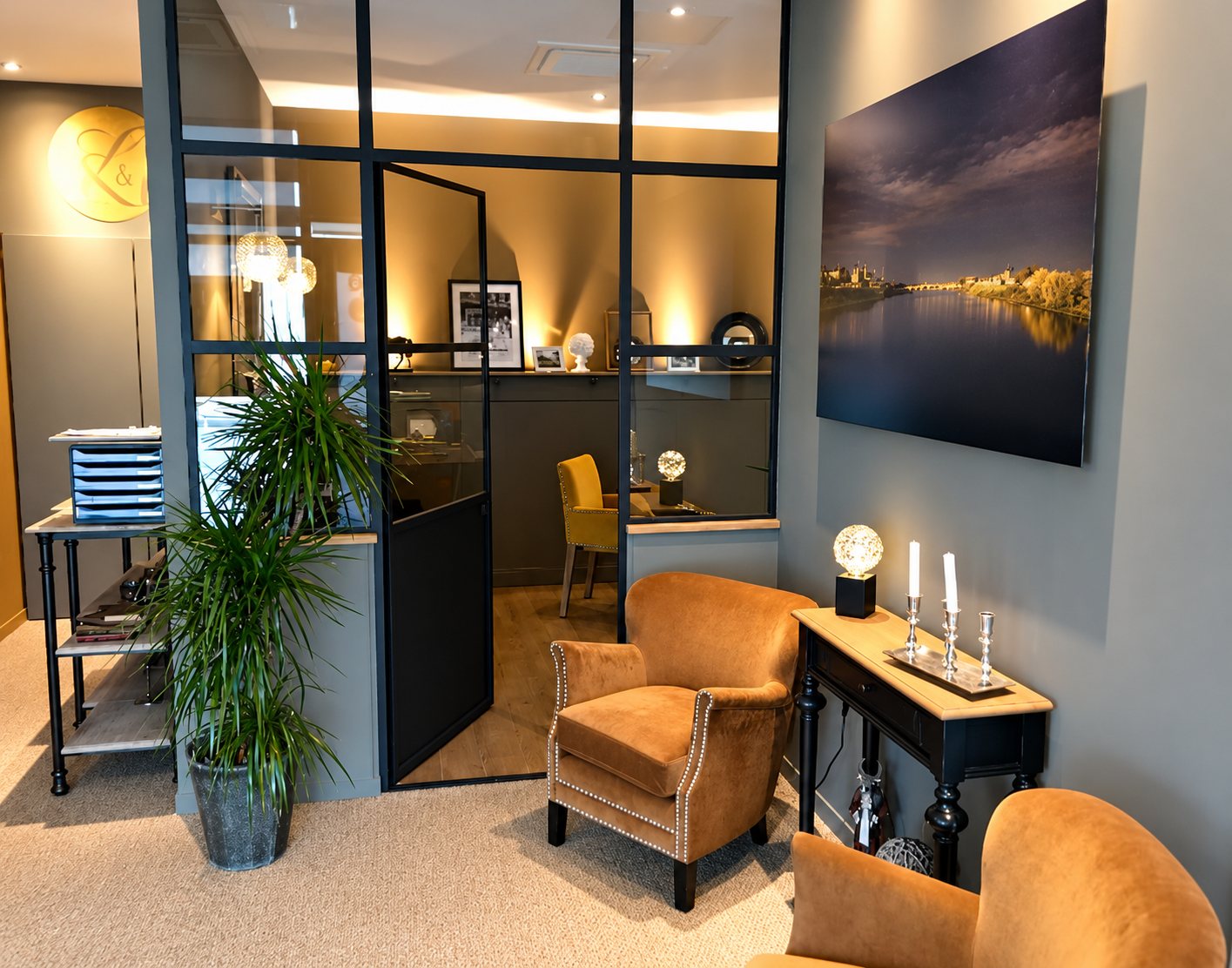 Espace d'accueil chaleureux avec deux fauteuils en velours moutarde, une console en bois, une grande plante verte et une verrière d'atelier noire donnant sur un bureau.