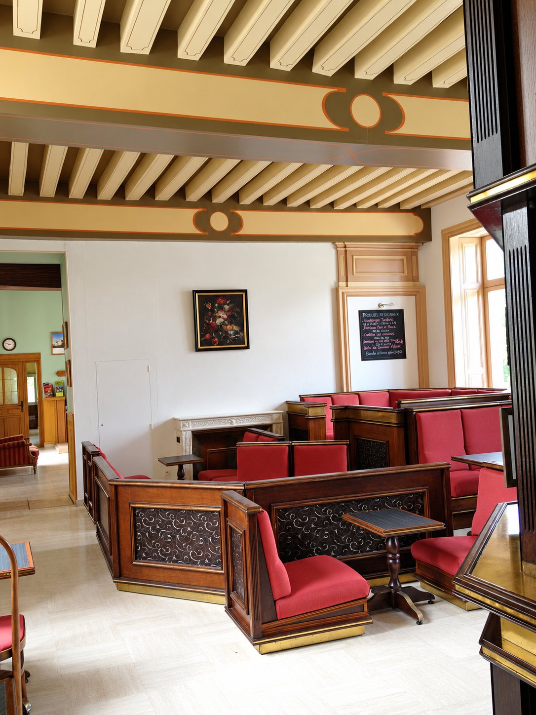 Salle élégante avec des banquettes en bois et velours rouge, de petites tables et un plafond à poutres peintes, avec une ardoise au mur du fond.