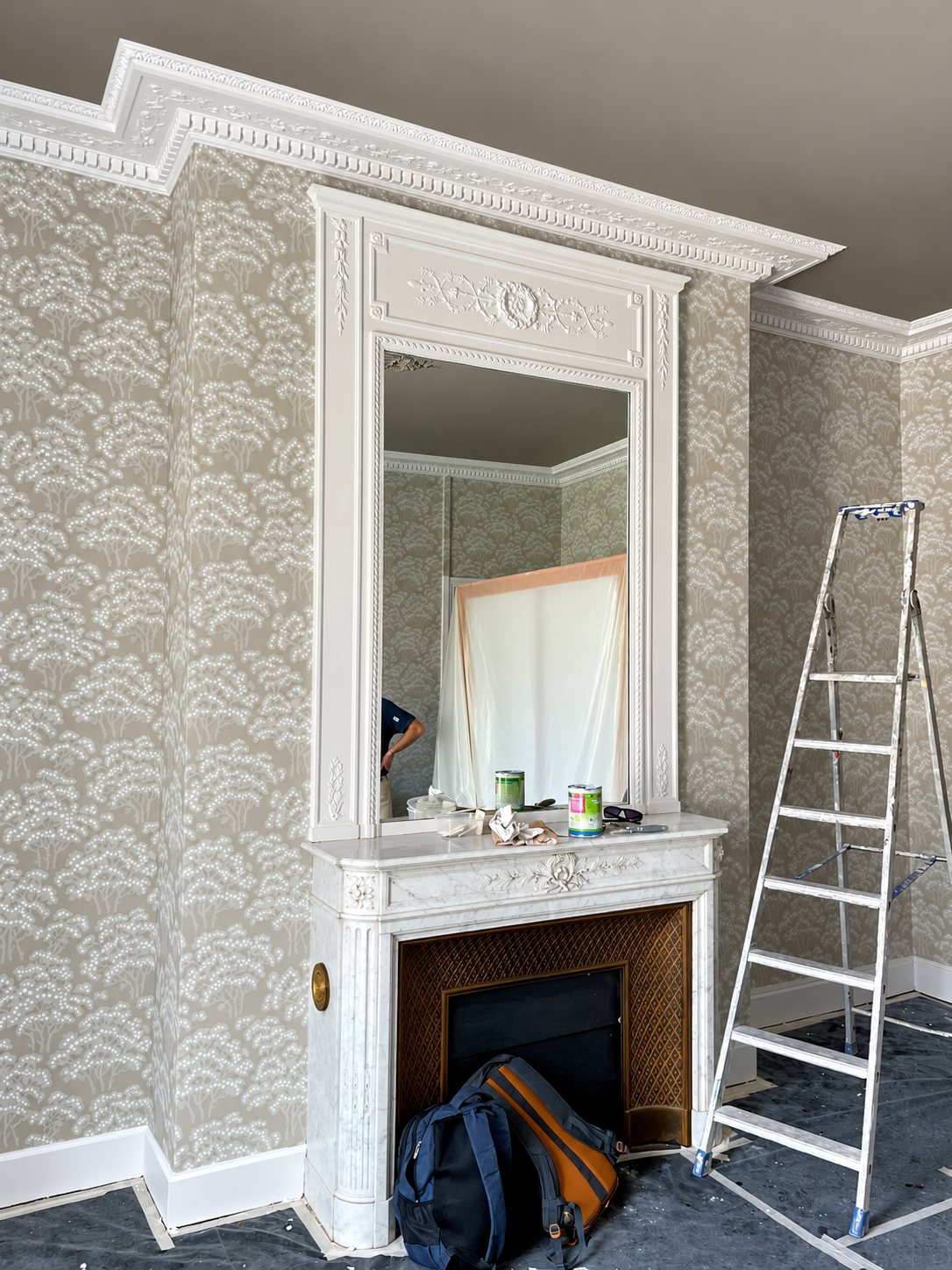 Pièce en cours de rénovation avec papier peint beige à motifs d'arbres, cheminée en marbre blanc surmontée d'un grand miroir, échelle et bâches de protection au sol.