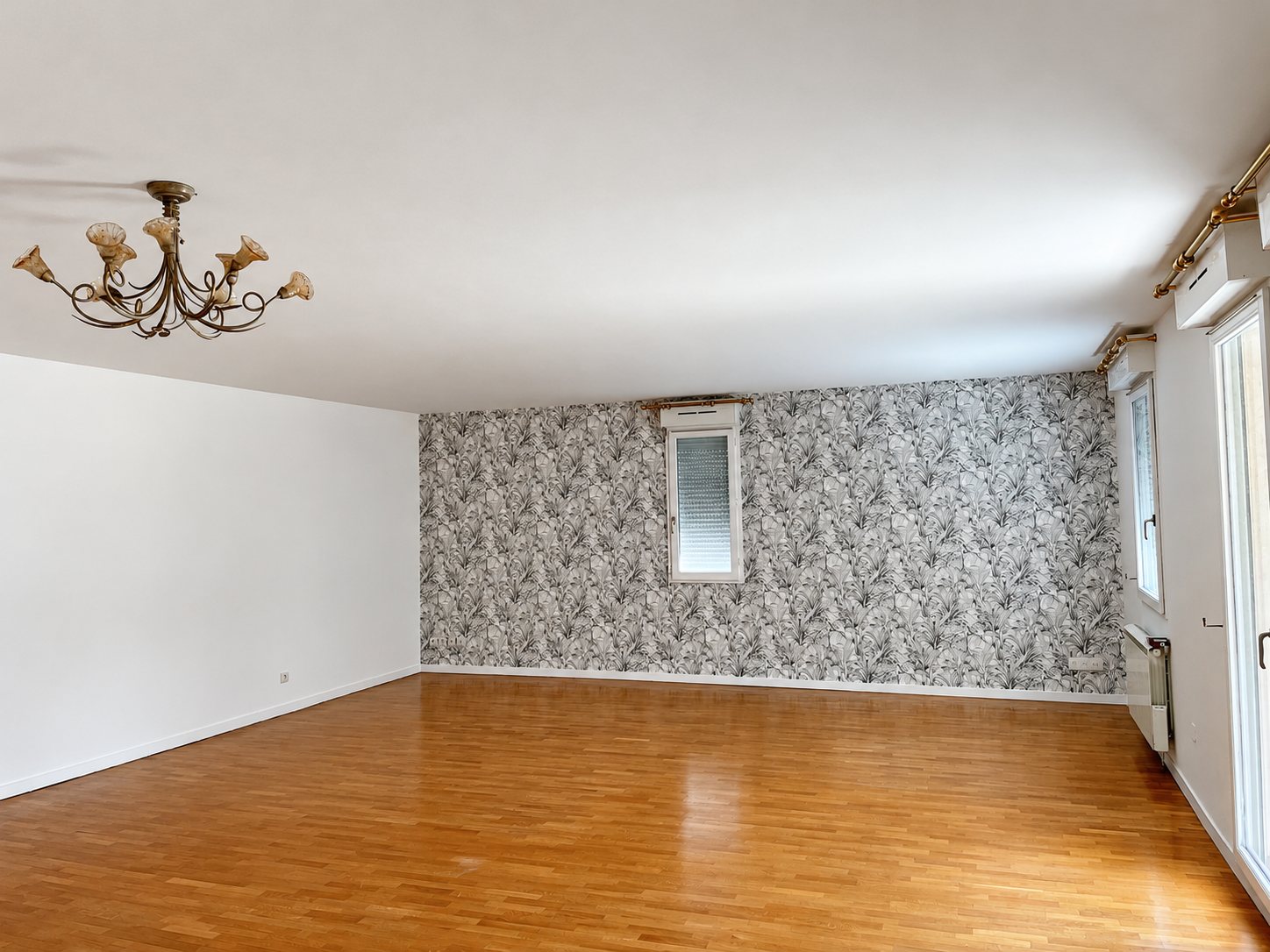 Grand salon vide au parquet clair, avec un mur d'accent en papier peint à motif de feuillages gris et un lustre ancien au plafond.