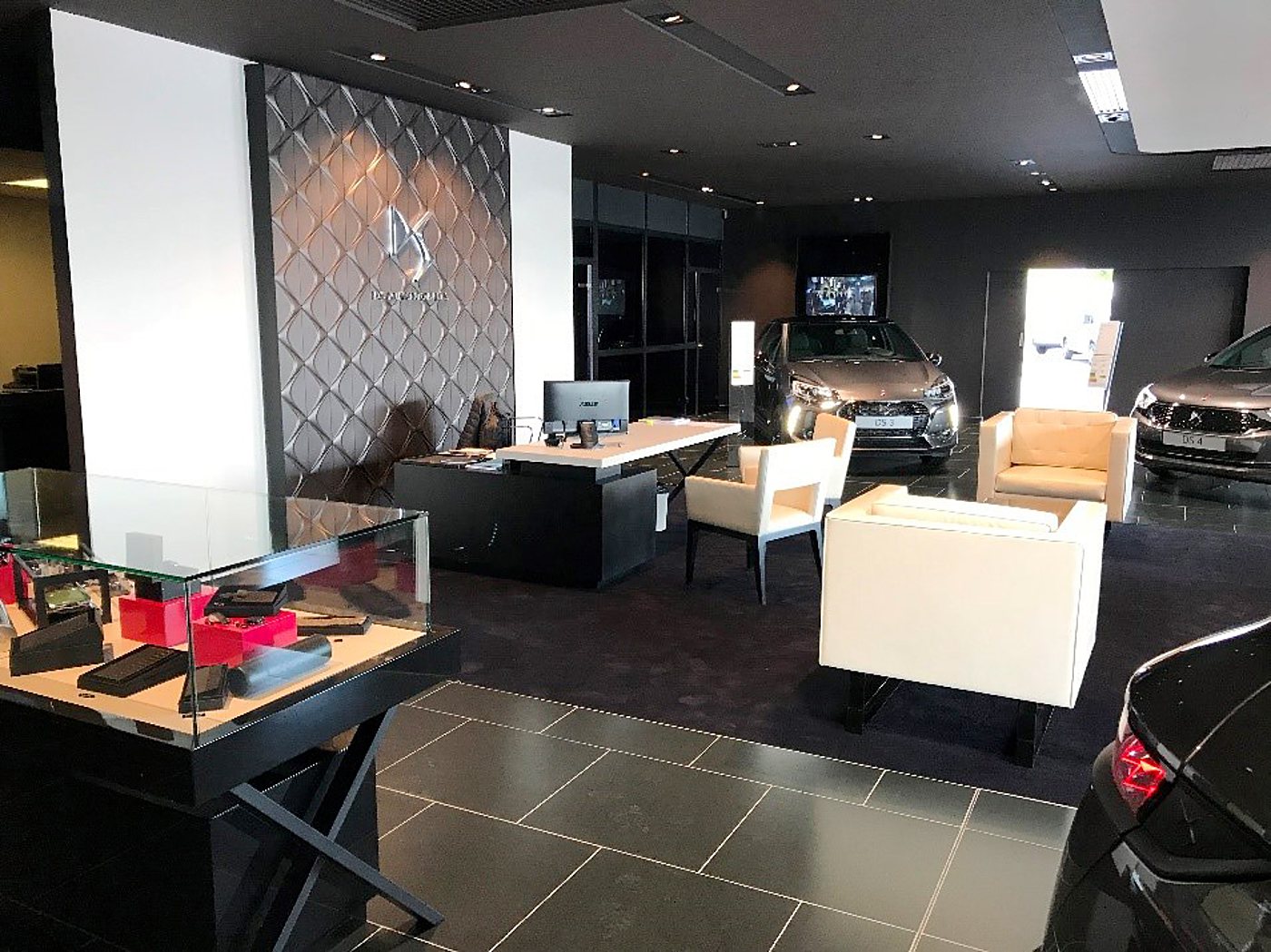 Intérieur d'un showroom automobile sombre et élégant avec un bureau d'accueil, des fauteuils crème, une vitrine d'accessoires et plusieurs voitures exposées.