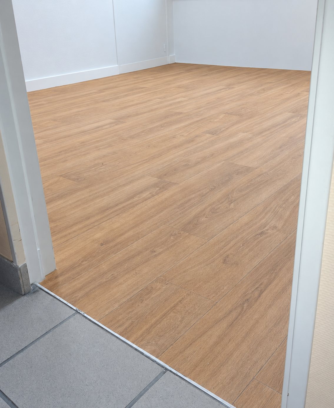 Une pièce vide aux murs blancs dont le sol est recouvert d'un revêtement imitation parquet clair, vu depuis une porte donnant sur un carrelage gris.